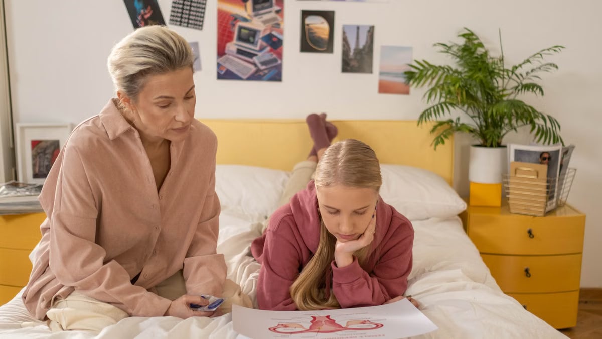 A mother and daughter talk over illustrated materials in a cozy bedroom setting.